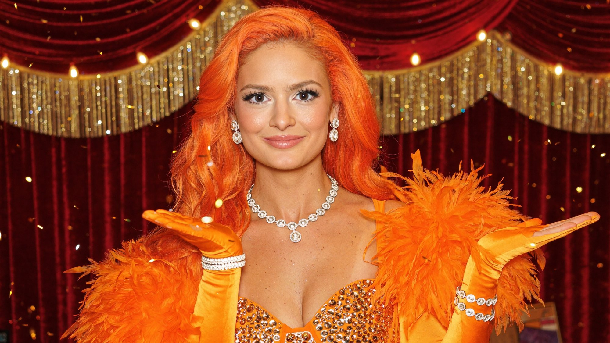 Orange-haired Carla in orange feathers and a diamond necklace blowing a kiss on a red-and-gold stage at the finale.