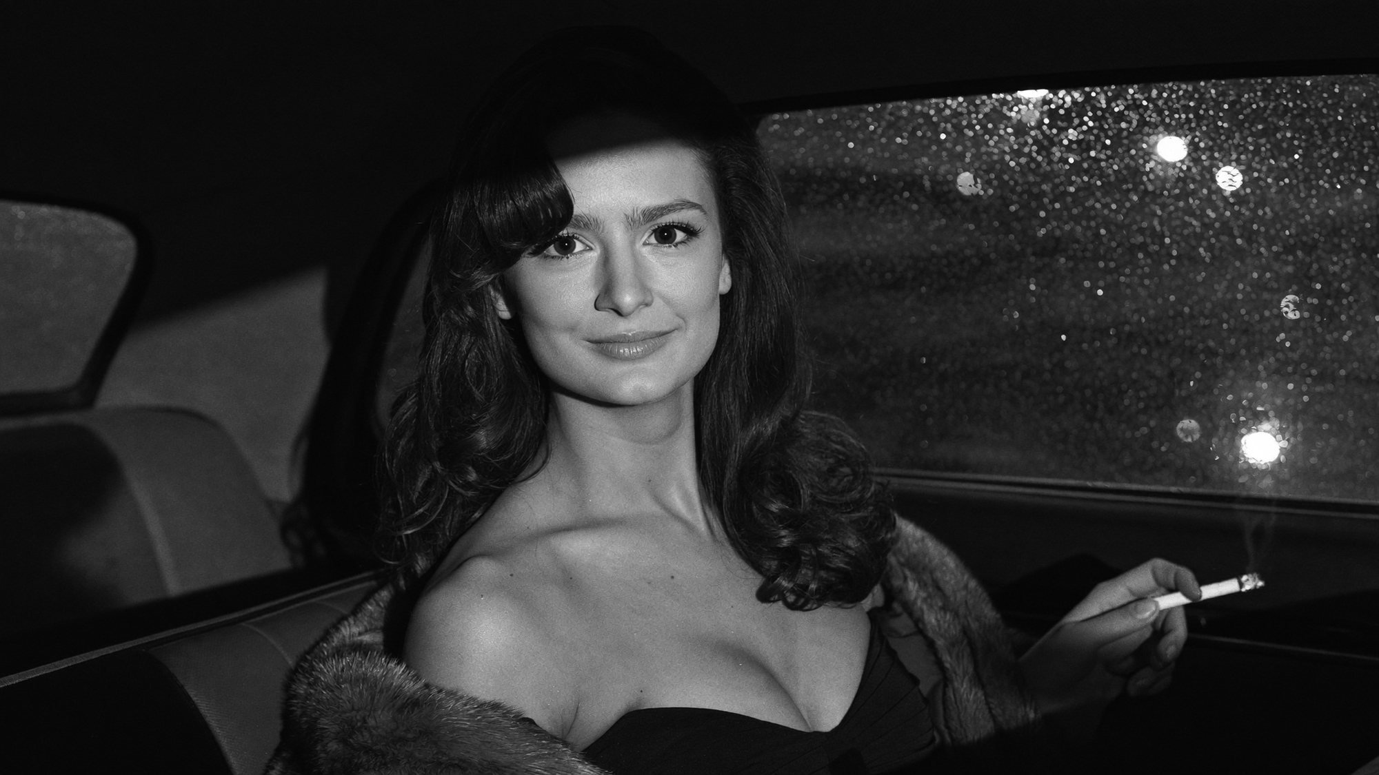 Carla in a fur-collared coat in the back seat of a sedan at night, cigarette in hand, rain on the window. Black and white.